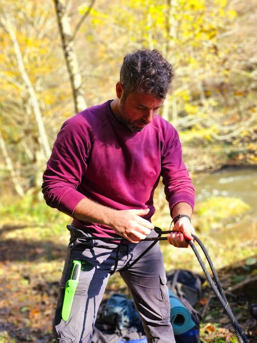 Man practicing rope skills during a survival and bushcraft course in the forest, with knife and gear visible, surrounded by autumn foliage near a river.