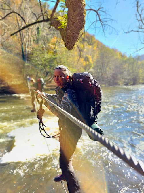 Woman crossing a river on a rope bridge during a survival and bushcraft course, wearing a large backpack and outdoor gear in a forest setting.