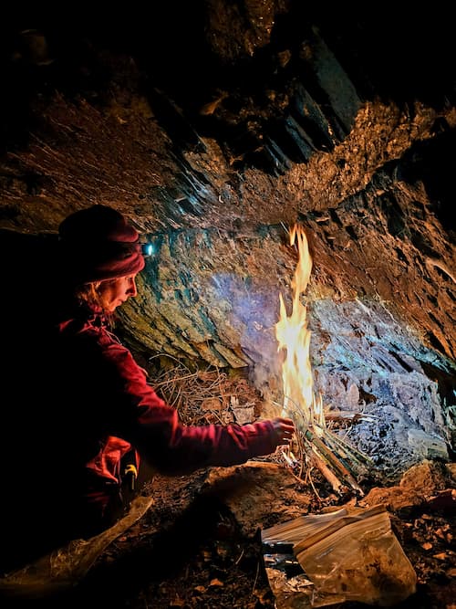 Person starting a fire at night under a rocky overhang during a survival and bushcraft course, using natural materials and a headlamp for visibility.