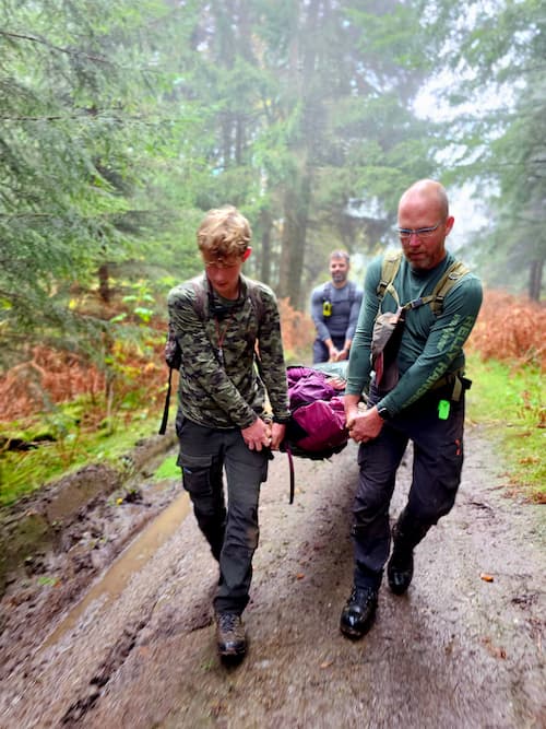 Three men carry a stretcher through a muddy forest trail during a survival exercise.