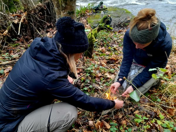 2 personen steken een beginnend vuurtje op de bosgrond